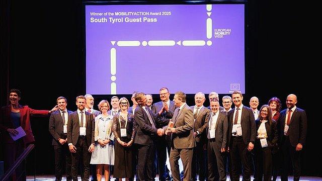 © LPA/Ivan Brentgani Die Südtiroler Delegation steht auf der Bühne, und einige ihrer Mitglieder applaudieren. In der Mitte der Delegation steht der Landesrat für Mobilität, Daniel Alfreider, der eine Trophäe in der Hand hält. Hinter ihm befindet sich eine Leinwand, auf der „Winner of the MOBILITYACTION Award 2025 South Tyrol Guest Pass“ steht.
