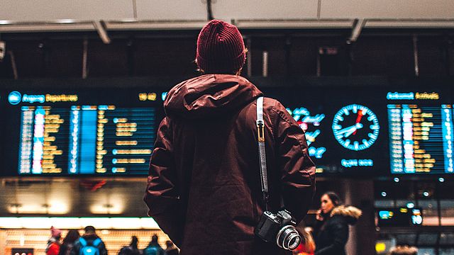 - Ragazzo di spalle con cappelli e macchina fotografica sulla spalla guarda il tabellone delle partenze della stazione dei treni.