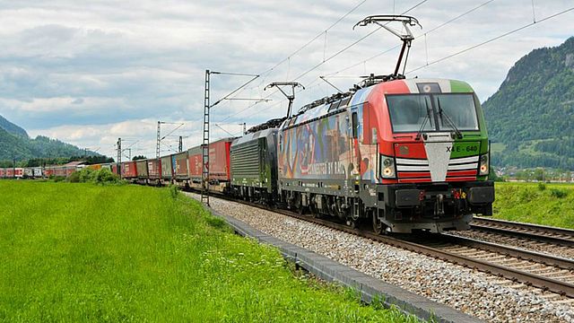 Ein Güterzug in einem Tal welches auf beiden Seiten von Bergen flankiert wird