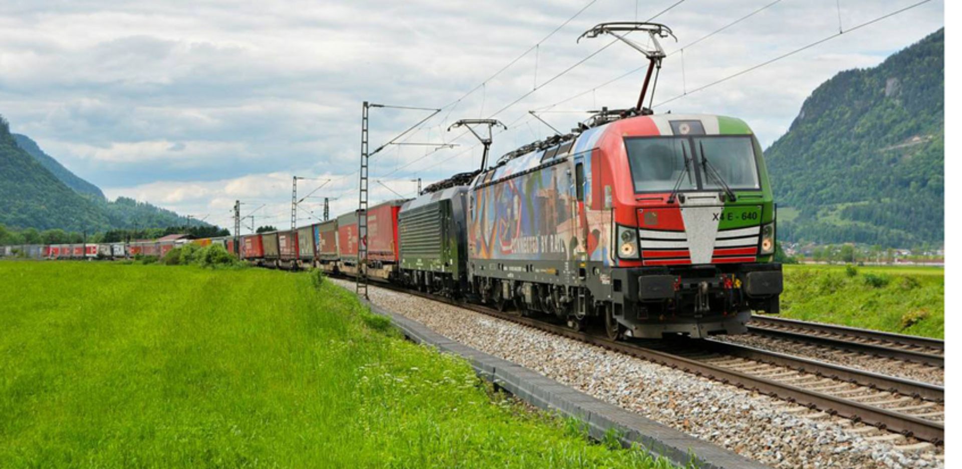 Ein Güterzug in einem Tal welches auf beiden Seiten von Bergen flankiert wird