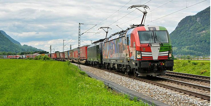 Ein Güterzug in einem Tal welches auf beiden Seiten von Bergen flankiert wird