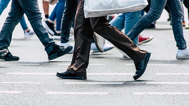 In primo piano, un uomo, di cui vediamo solo le gambe, attraversa la strada. Sullo sfondo, ci sono diversi passanti. 